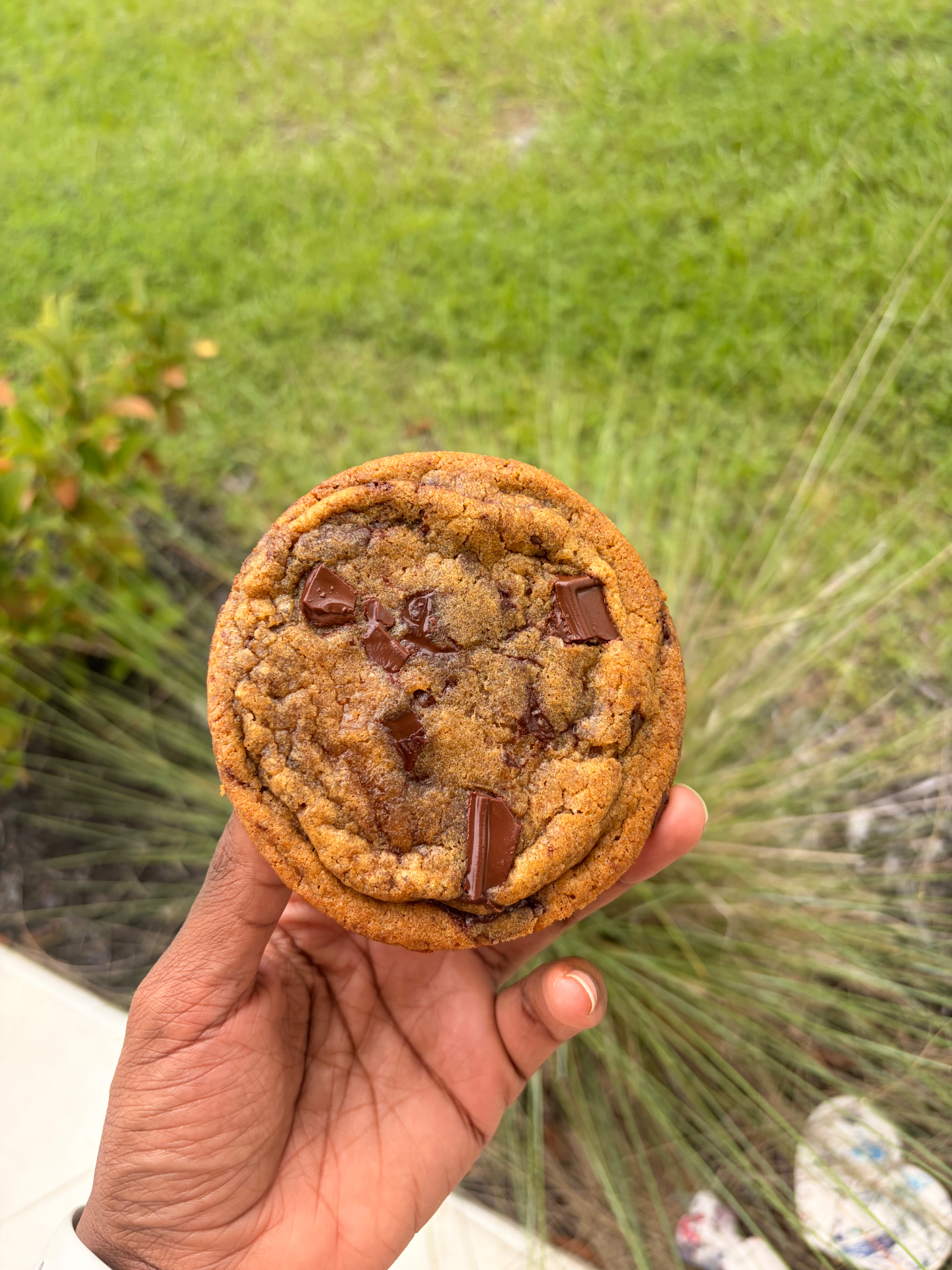 A single oversized chocolate chunk cookie with melting pools of dark chocolate, held in a hand above a green lawn.